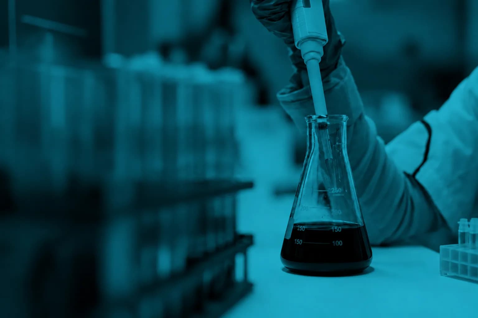 A gloved hand pipetting into a conical flask on a laboratory bench